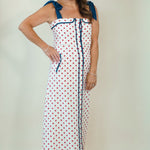 A woman modeling a sleeveless white maxi dress with red polka dots and navy blue trim, featuring shoulder ties and a front tie closure, set against a plain cream studio background.