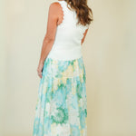 A woman shown from the back wearing a sleeveless white top with scalloped edges and a long, tiered maxi skirt featuring a light blue, green, and white floral and gingham pattern, set against a plain beige studio background.