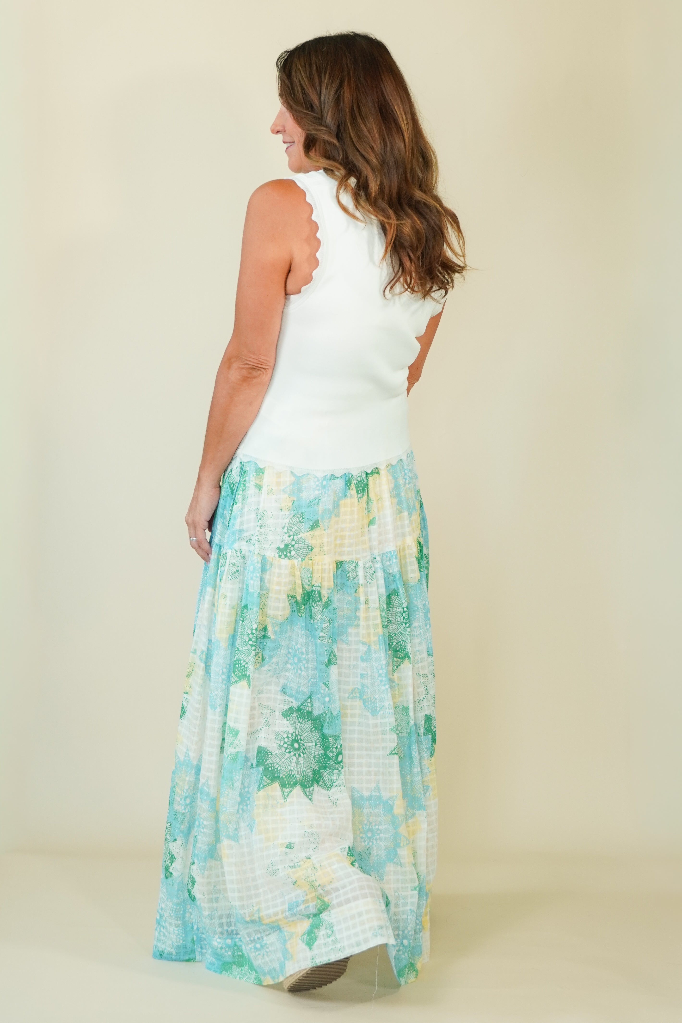 A woman shown from the back wearing a sleeveless white top with scalloped edges and a long, tiered maxi skirt featuring a light blue, green, and white floral and gingham pattern, set against a plain beige studio background.