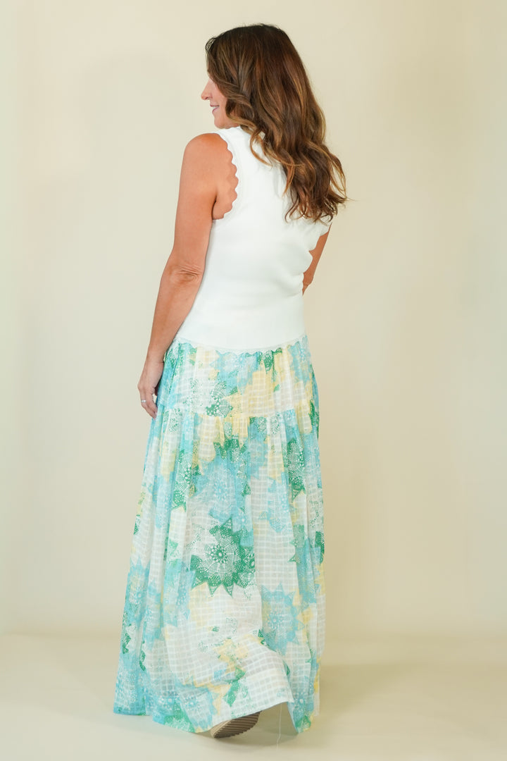A woman shown from the back wearing a sleeveless white top with scalloped edges and a long, tiered maxi skirt featuring a light blue, green, and white floral and gingham pattern, set against a plain beige studio background.