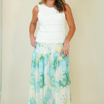 A woman modeling a sleeveless white top paired with a long, flowing maxi skirt featuring a watercolor-style floral pattern in shades of green, blue, and yellow on a white base, set against a plain beige studio background.