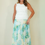 A woman modeling a sleeveless white top and a long, flowing maxi skirt with a light blue, green, and yellow floral pattern, set against a plain beige studio background.