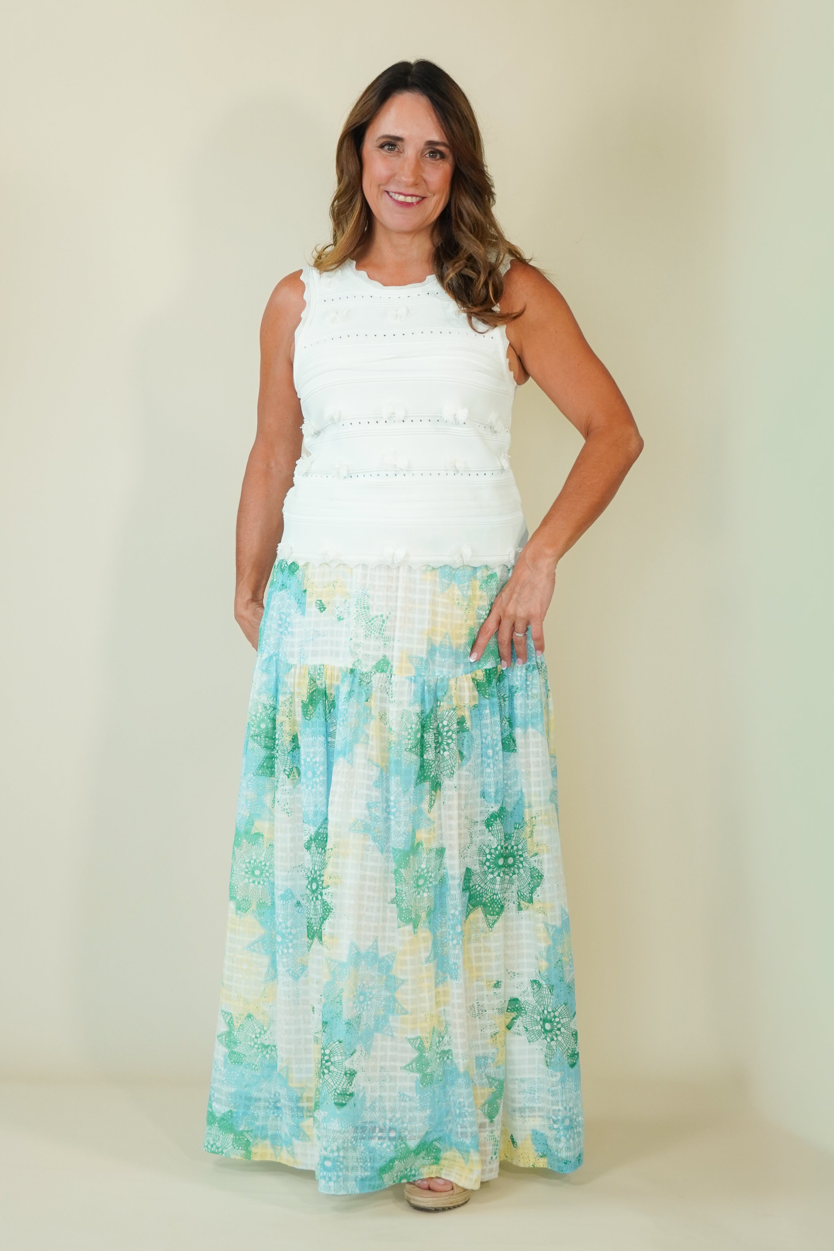 A woman modeling a sleeveless white top and a long, flowing maxi skirt with a light blue, green, and yellow floral pattern, set against a plain beige studio background.