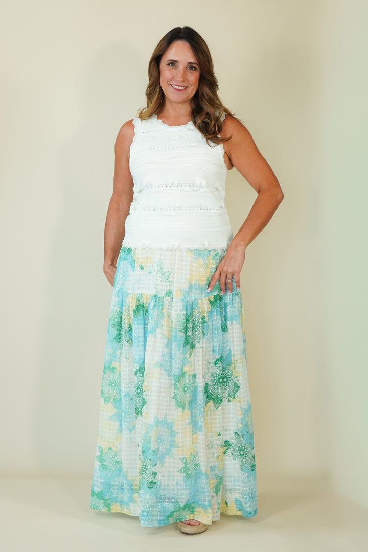 A woman modeling a sleeveless white top and a long, flowing maxi skirt with a light blue, green, and yellow floral pattern, set against a plain beige studio background.
