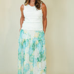 A woman modeling a sleeveless white crop top and a long tiered maxi skirt with a blue, green, and white watercolor floral pattern, standing against a plain beige background.