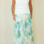 A woman modeling a sleeveless maxi dress with a white textured bodice and a tiered skirt featuring a green, blue, and yellow floral pattern on a white base, set against a plain beige studio background.