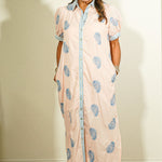 Woman wearing a long, light pink dress with blue floral patterns against a plain wall.