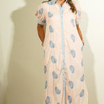 Woman wearing a long, light-colored dress with blue floral patterns against a plain wall.