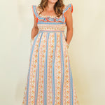 A woman modeling a sleeveless maxi dress with a cream base, vertical blue and orange stripes, and a smocked bodice, standing against a plain cream background.