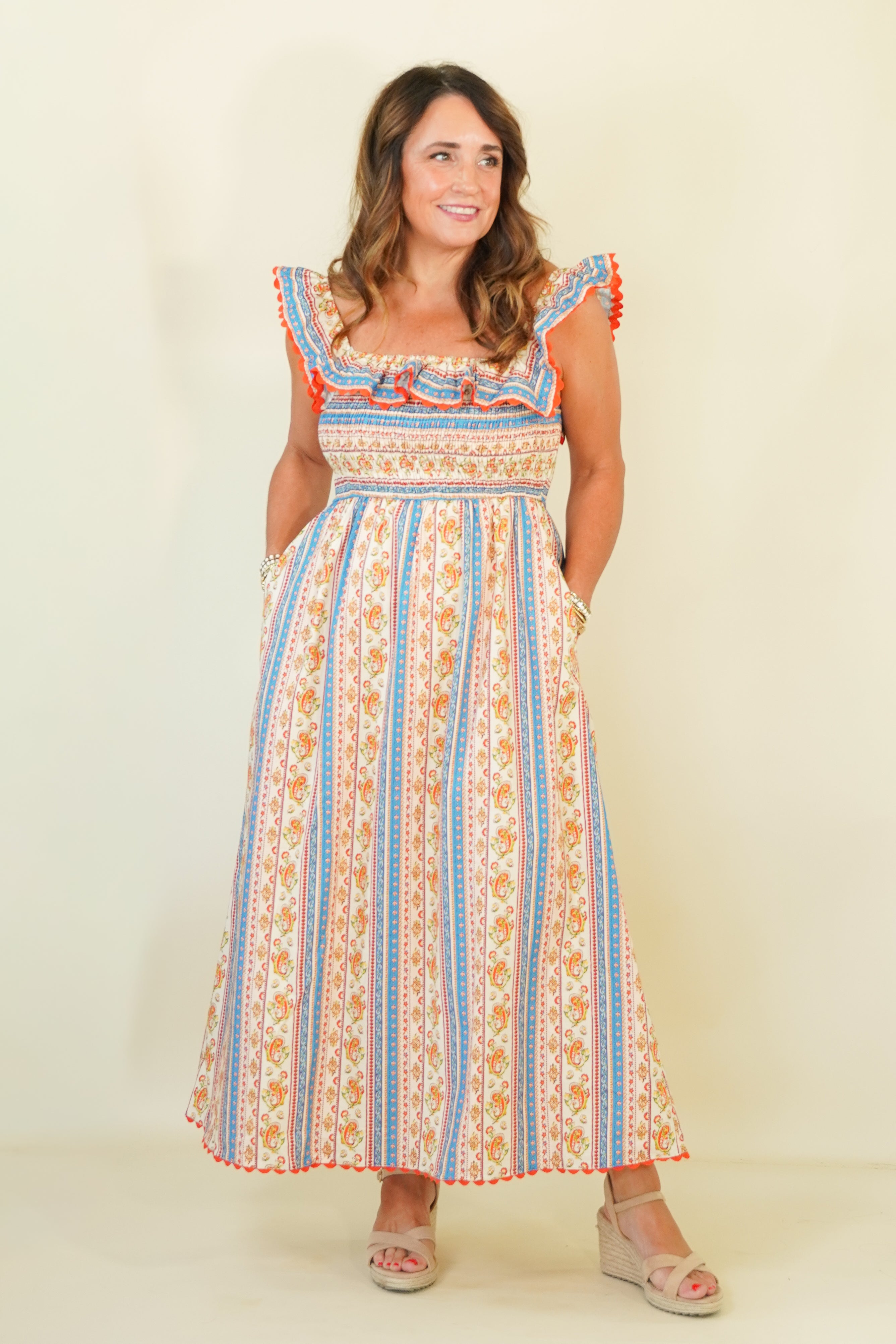 A woman modeling a sleeveless maxi dress with a cream base, vertical blue and orange stripes, and a smocked bodice, standing against a plain cream background.