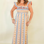 A woman modeling a sleeveless maxi dress with a colorful vertical stripe pattern in orange, blue, and cream, featuring ruffled shoulder straps and a smocked bodice, set against a plain cream background.