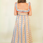 A woman viewed from the back wearing a sleeveless maxi dress with ruffled shoulder straps, a square neckline, and a vertical striped pattern in orange, blue, and cream. She is standing against a plain cream background.