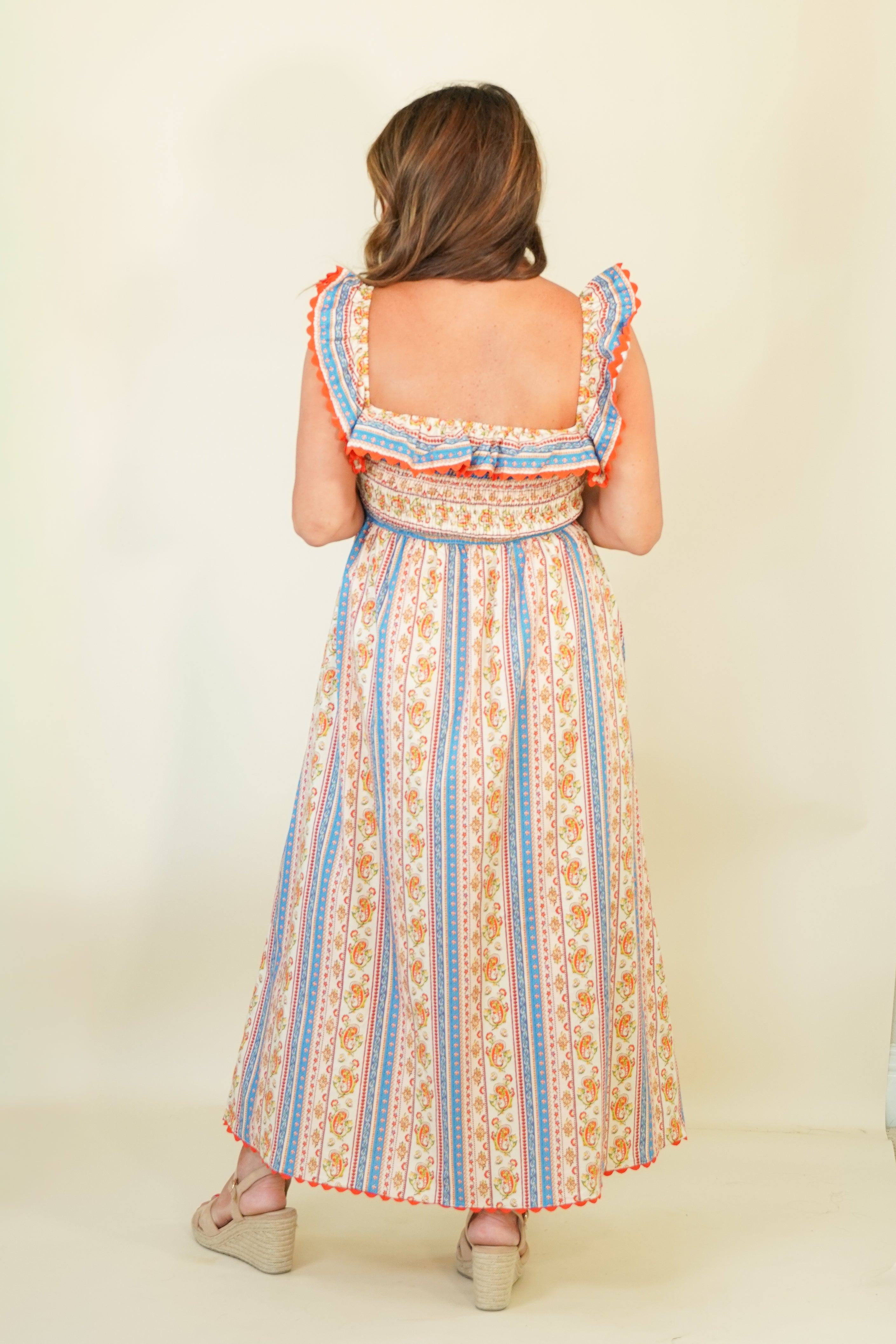 A woman viewed from the back wearing a sleeveless maxi dress with ruffled shoulder straps, a square neckline, and a vertical striped pattern in orange, blue, and cream. She is standing against a plain cream background.