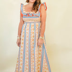 A woman modeling a bohemian-style maxi dress with vertical stripes in blue, orange, and cream, featuring ruffled shoulder straps and a smocked bodice, set against a plain cream background.