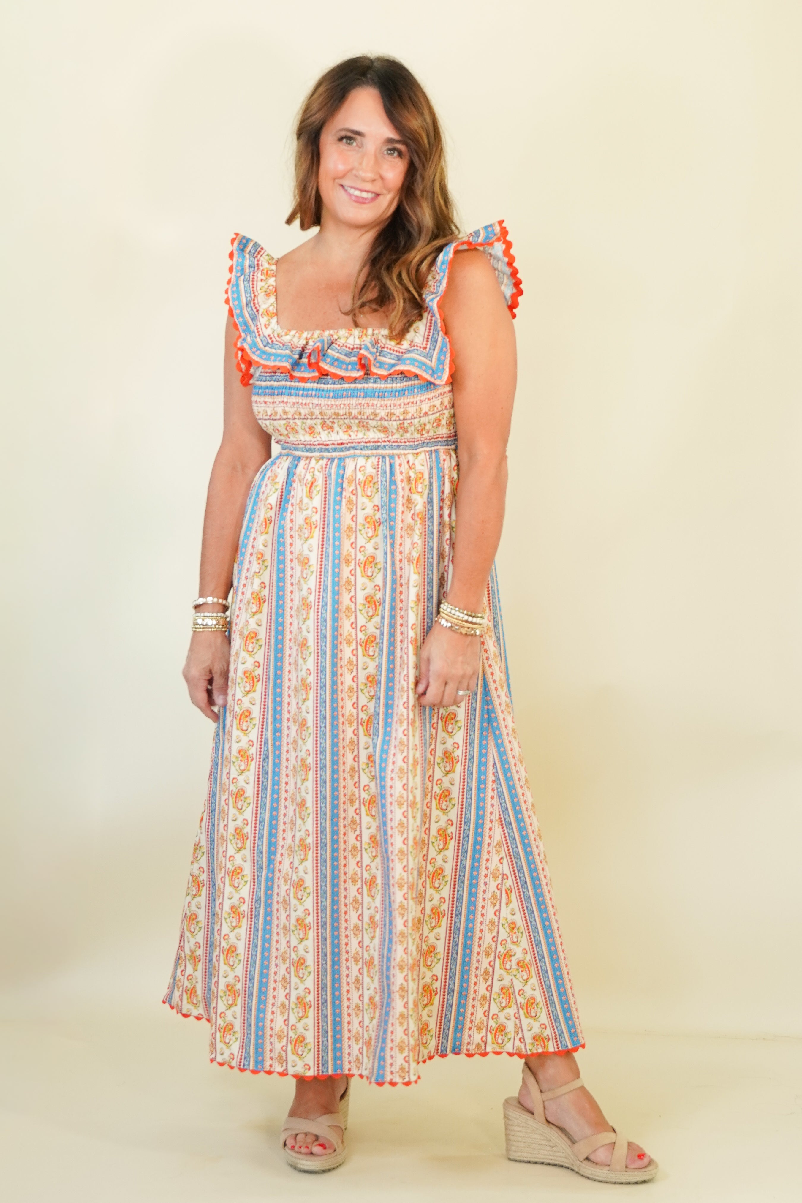 A woman modeling a bohemian-style maxi dress with vertical stripes in blue, orange, and cream, featuring ruffled shoulder straps and a smocked bodice, set against a plain cream background.