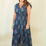 A woman modeling a sleeveless navy blue maxi dress with a pink and green floral pattern, standing against a plain cream background.
