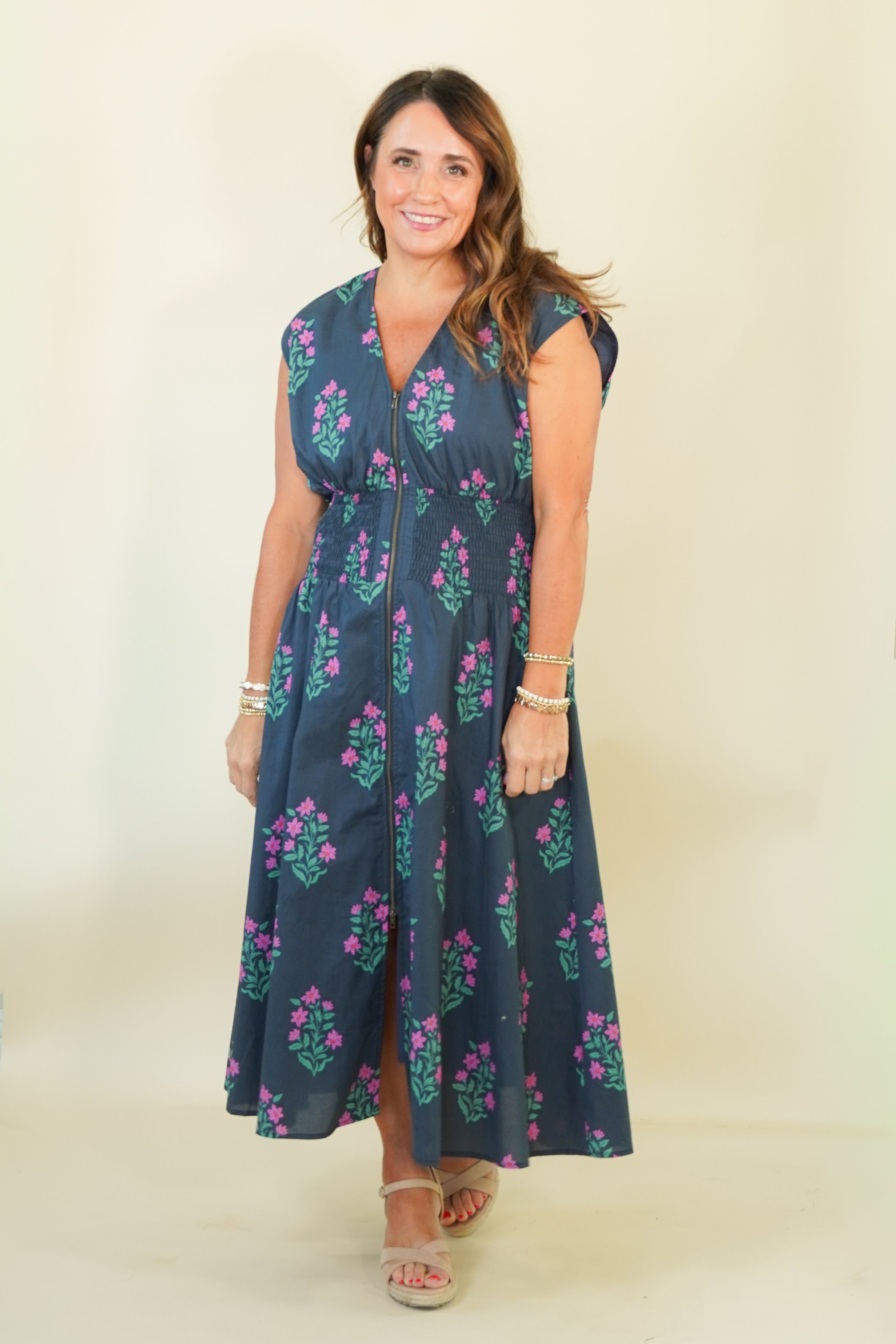 A woman modeling a sleeveless navy blue maxi dress with a pink and green floral pattern, standing against a plain cream background.