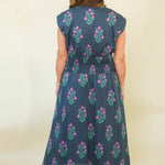 A woman viewed from the back wearing a sleeveless midi dress with a dark blue base and pink floral block-print pattern, paired with beige wedge sandals against a plain cream background.