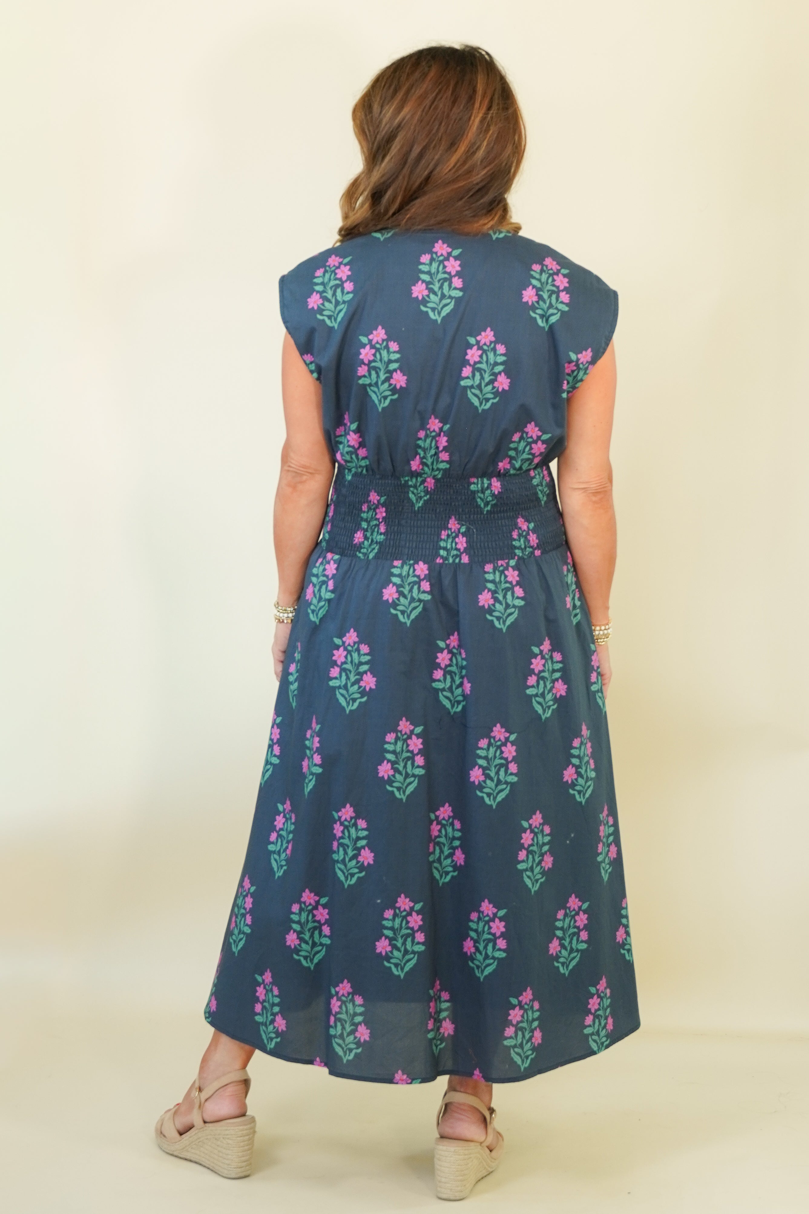 A woman viewed from the back wearing a sleeveless midi dress with a dark blue base and pink floral block-print pattern, paired with beige wedge sandals against a plain cream background.