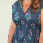 A woman modeling a sleeveless navy blue maxi dress with a pink and green floral pattern, standing against a plain cream-colored studio background.
