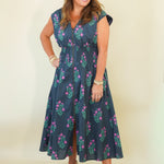 A woman modeling a sleeveless navy blue midi dress with a pink and green floral pattern, standing against a plain cream-colored background.