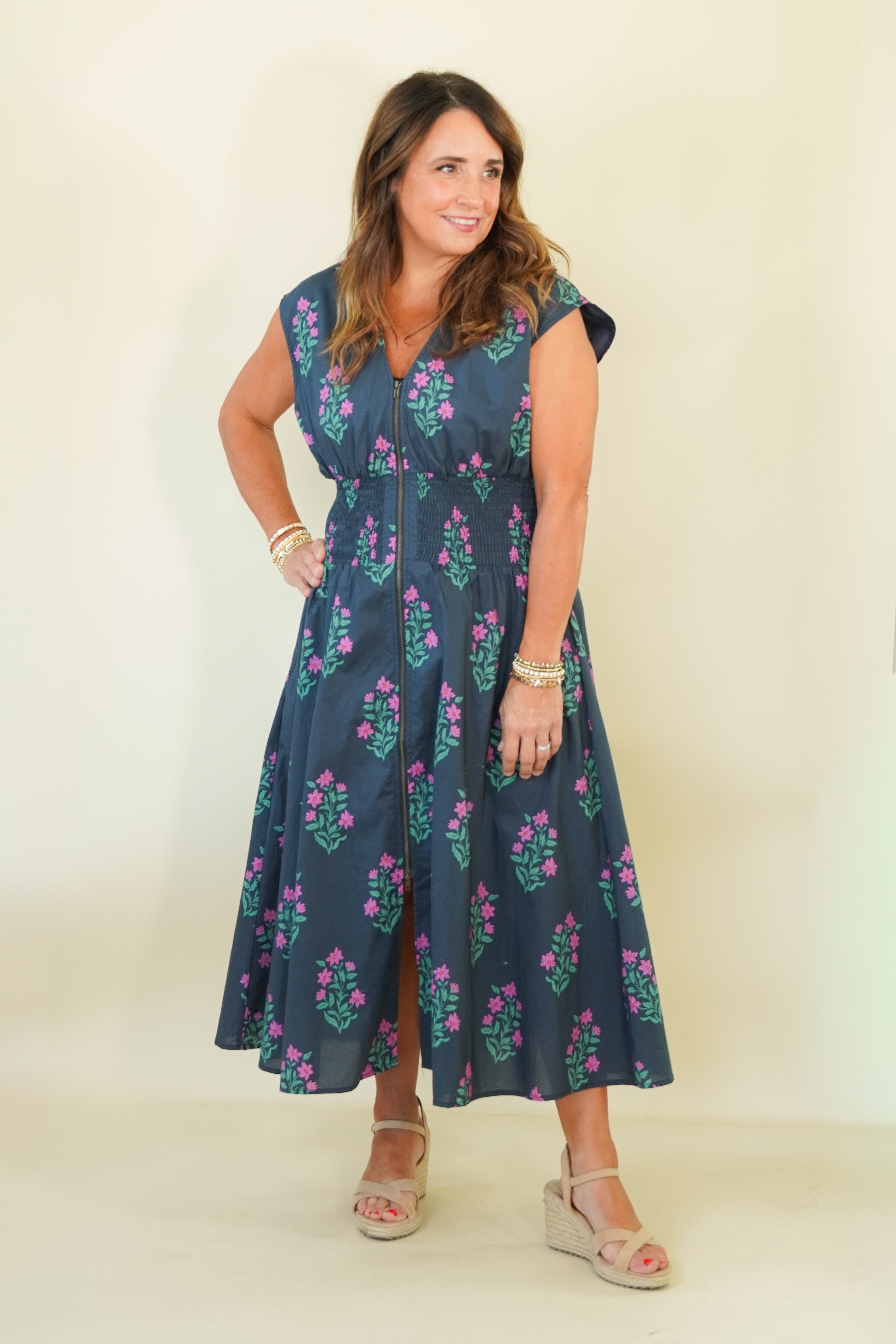 A woman modeling a sleeveless navy blue midi dress with a pink and green floral pattern, standing against a plain cream-colored background.