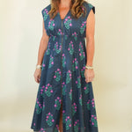 A woman modeling a sleeveless navy blue midi dress with a green and pink floral pattern, standing against a plain cream background.