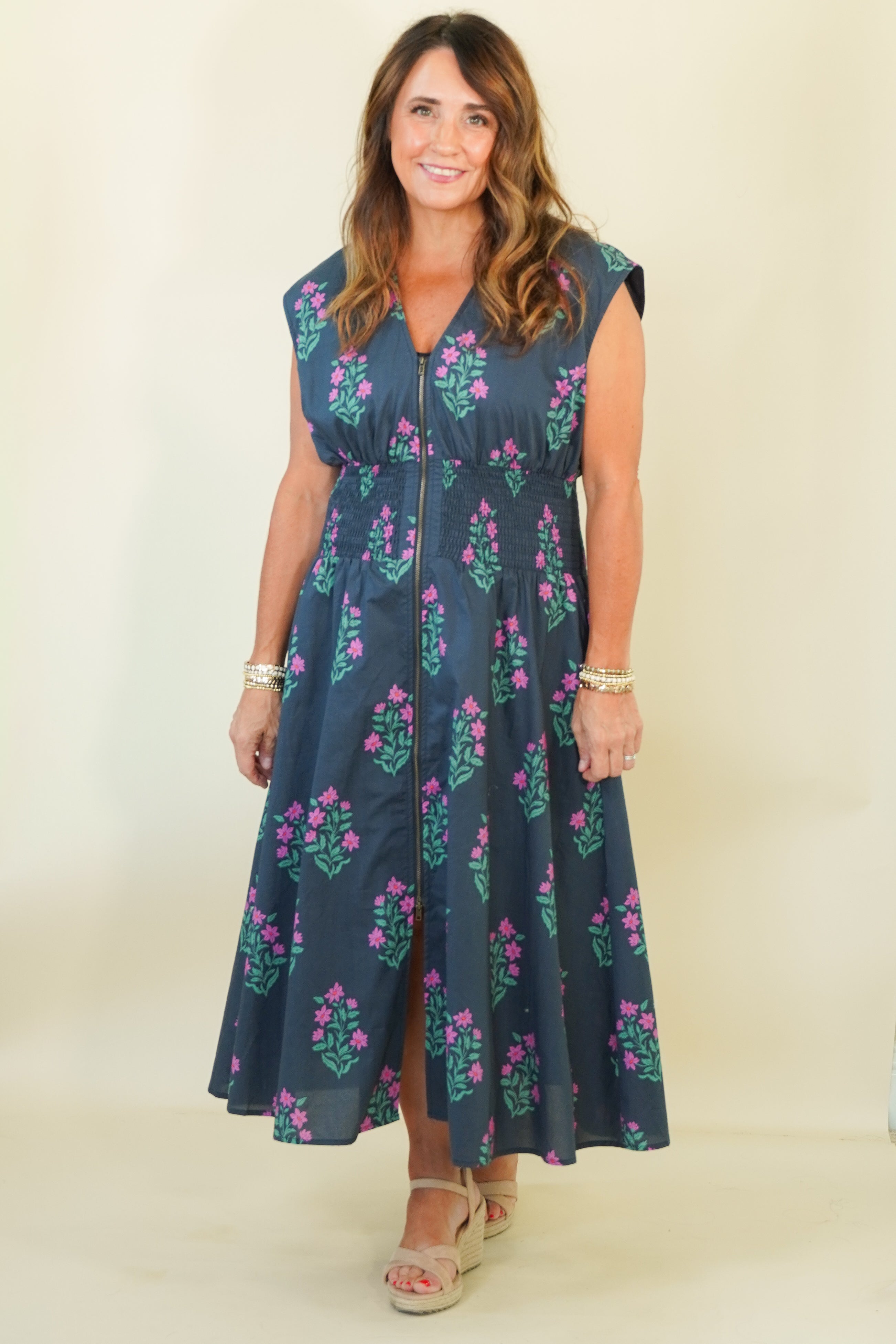 A woman modeling a sleeveless navy blue midi dress with a green and pink floral pattern, standing against a plain cream background.