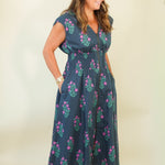A woman modeling a navy blue midi dress with a pink and green floral pattern, standing against a plain cream background.