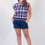 A woman modeling a sleeveless knit top with a navy, white, and red plaid pattern, paired with dark blue denim shorts and beige slide sandals, against a plain white studio background.