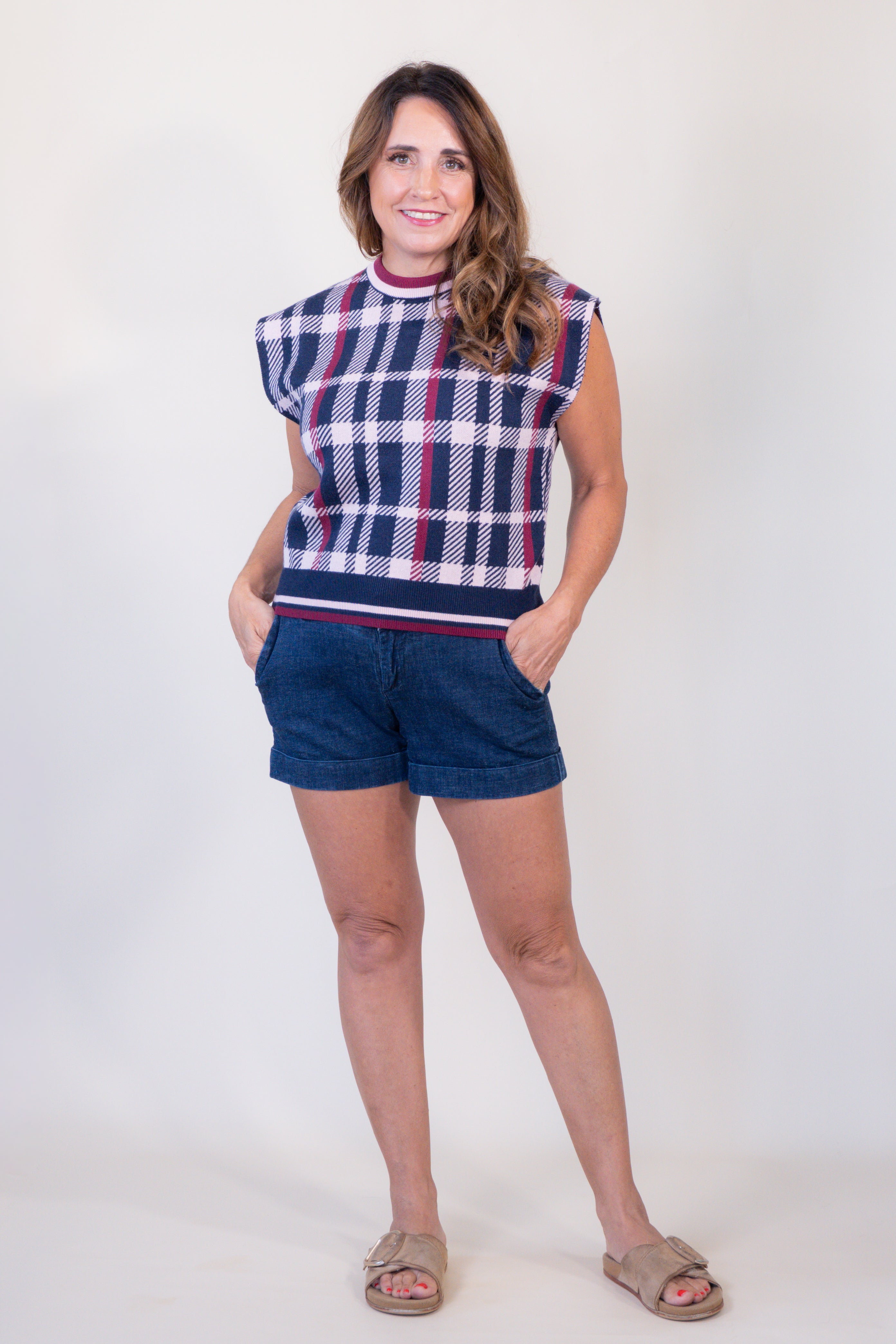A woman modeling a sleeveless knit top with a navy, white, and red plaid pattern, paired with dark blue denim shorts and beige slide sandals, against a plain white studio background.