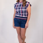A woman modeling a sleeveless knit top with a navy, white, and red plaid pattern, paired with dark blue denim shorts and tan sandals, against a plain light gray studio background.