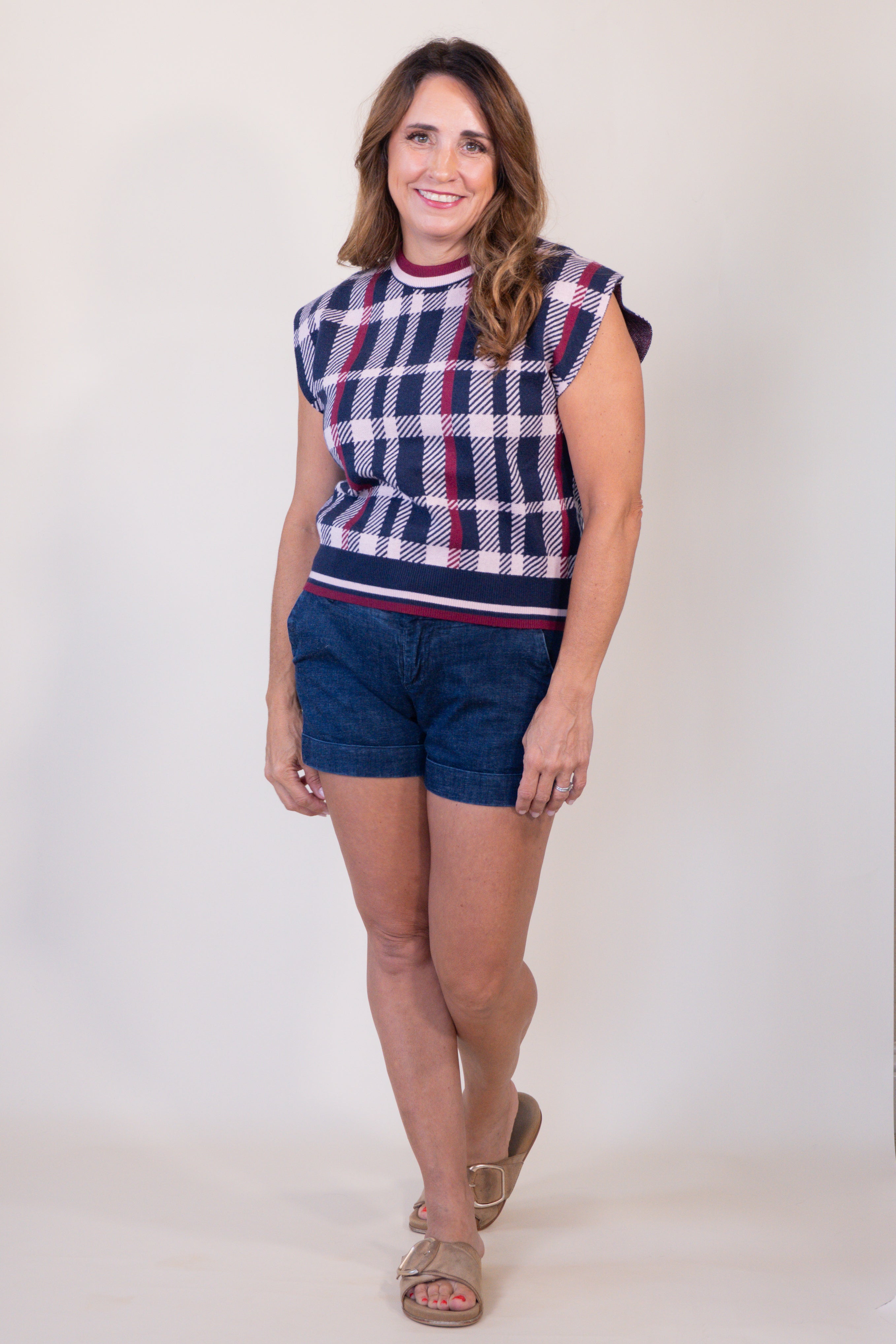 A woman modeling a sleeveless knit top with a navy, white, and red plaid pattern, paired with dark blue denim shorts and tan sandals, against a plain light gray studio background.