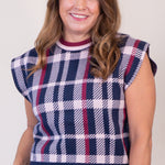 A smiling woman with long brown hair wearing a navy, white, and burgundy plaid sleeveless knit top with blue jeans, photographed against a plain light beige background.
