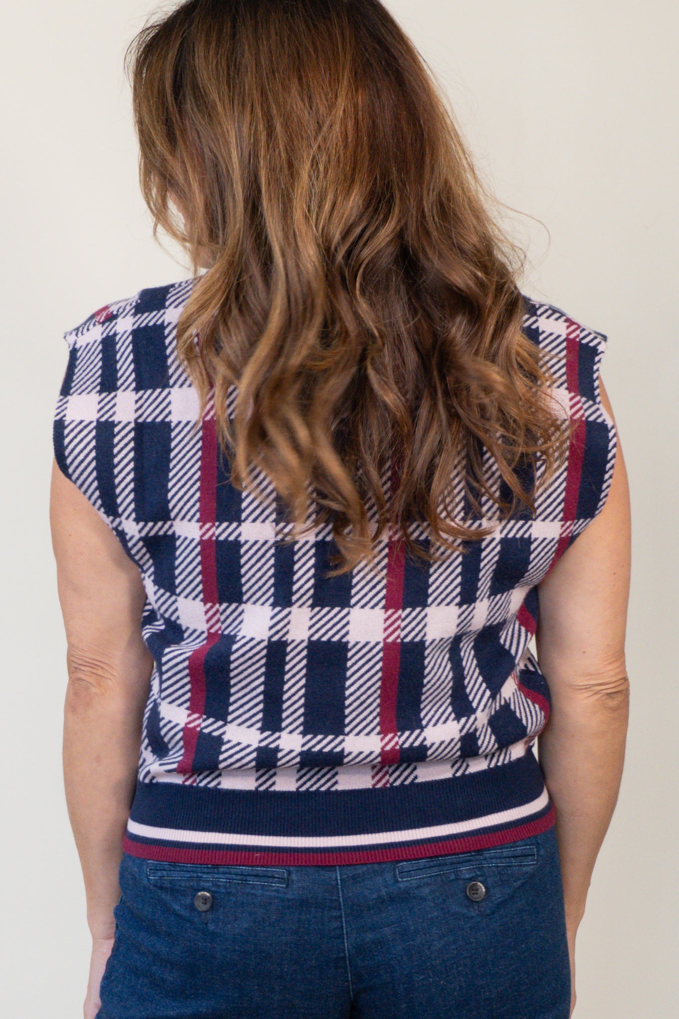 A woman viewed from behind wearing a navy, white, and red plaid sleeveless top with a striped hem, paired with blue jeans.