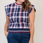 A woman modeling a sleeveless knit top with a navy, white, and red plaid pattern, paired with dark blue denim shorts and tan slide sandals, against a plain off-white studio background.
