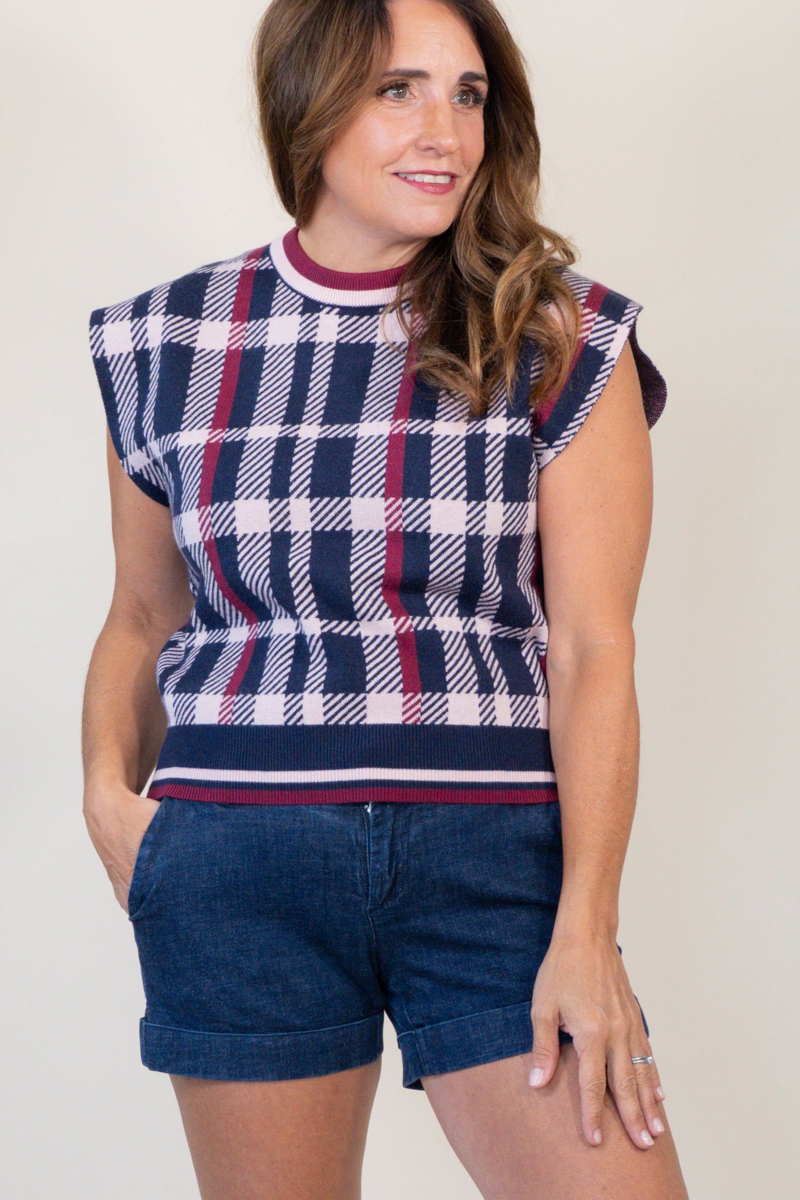 A woman modeling a sleeveless knit top with a navy, white, and red plaid pattern, paired with dark blue denim shorts and tan slide sandals, against a plain off-white studio background.