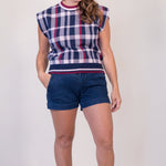 A woman modeling a sleeveless knit top with a navy, white, and red plaid pattern, paired with dark blue denim shorts and tan slide sandals, against a plain light beige studio background.