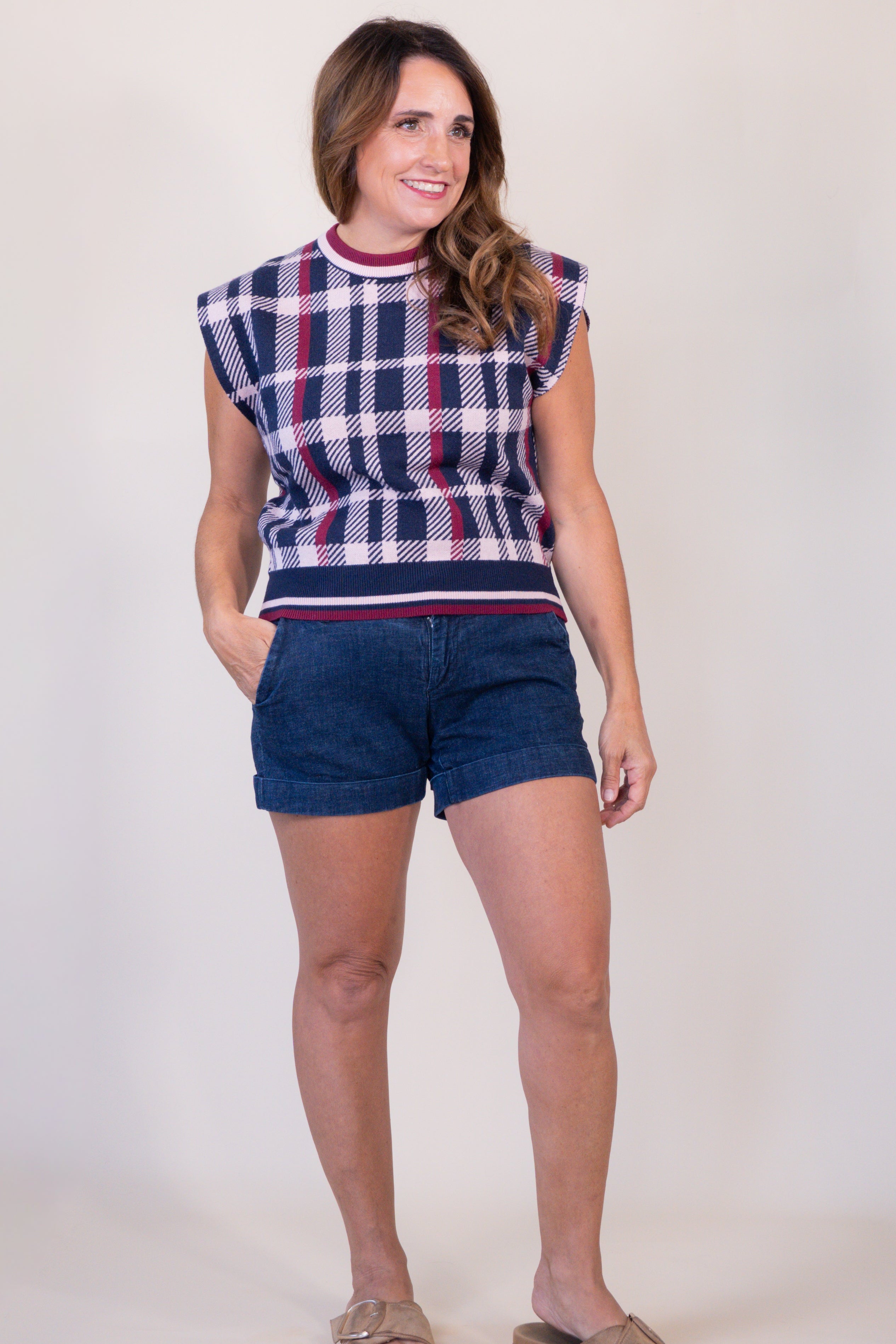A woman modeling a sleeveless knit top with a navy, white, and red plaid pattern, paired with dark blue denim shorts and tan slide sandals, against a plain light beige studio background.