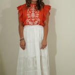Woman wearing a red blouse with white floral patterns and a white lace skirt against a plain background