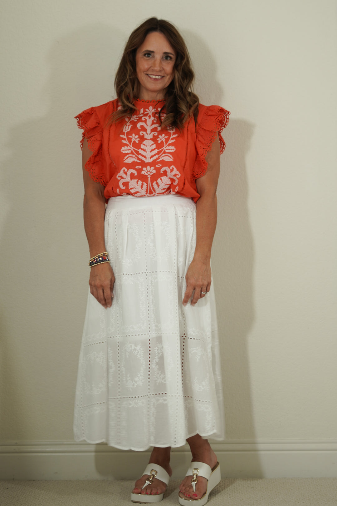 Woman wearing a red blouse with white floral patterns and a white lace skirt against a plain background