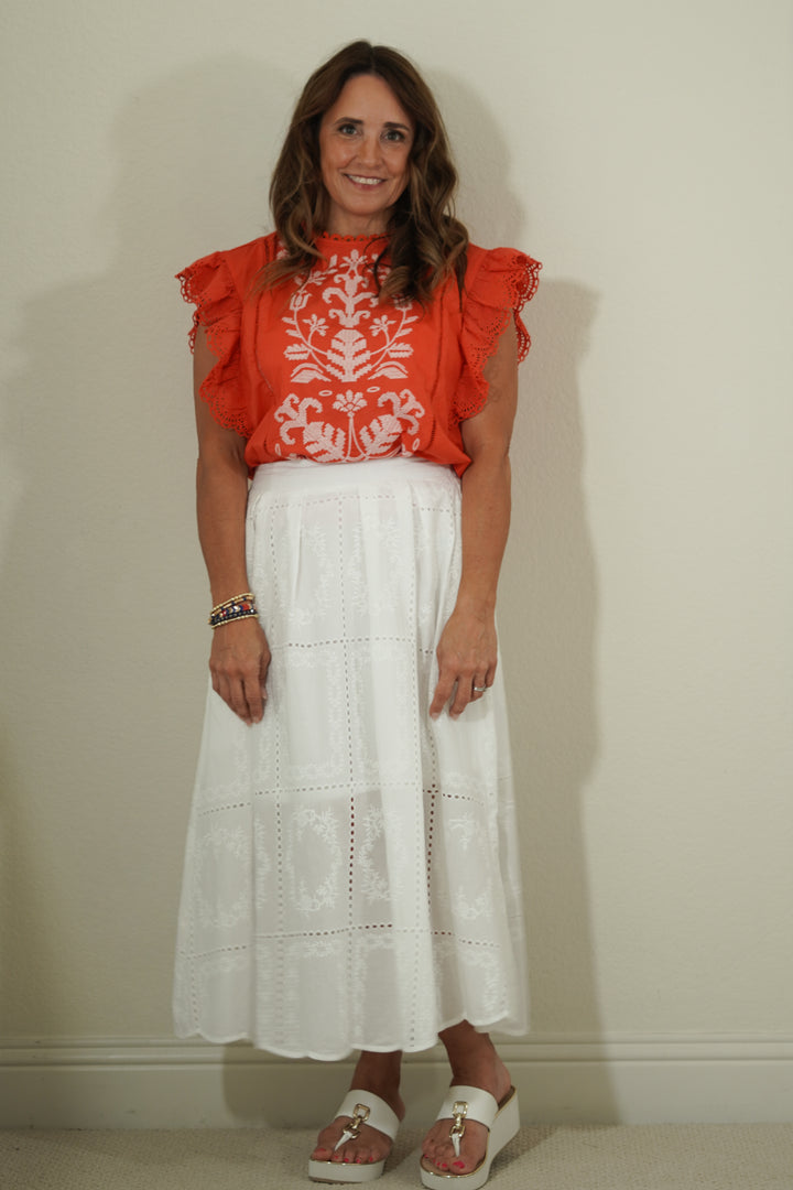 Woman wearing a red blouse with white floral patterns and a white lace skirt against a plain background