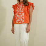 Woman wearing an orange top with white floral patterns and white pants against a beige wall.