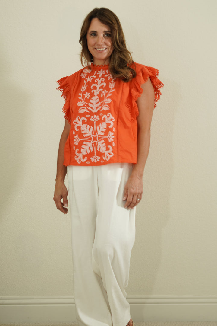 Woman wearing an orange top with white floral patterns and white pants against a beige wall.