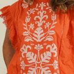 Person wearing an orange top with white floral embroidery against a plain background