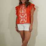 Woman wearing an orange top with white patterns and white shorts against a plain background