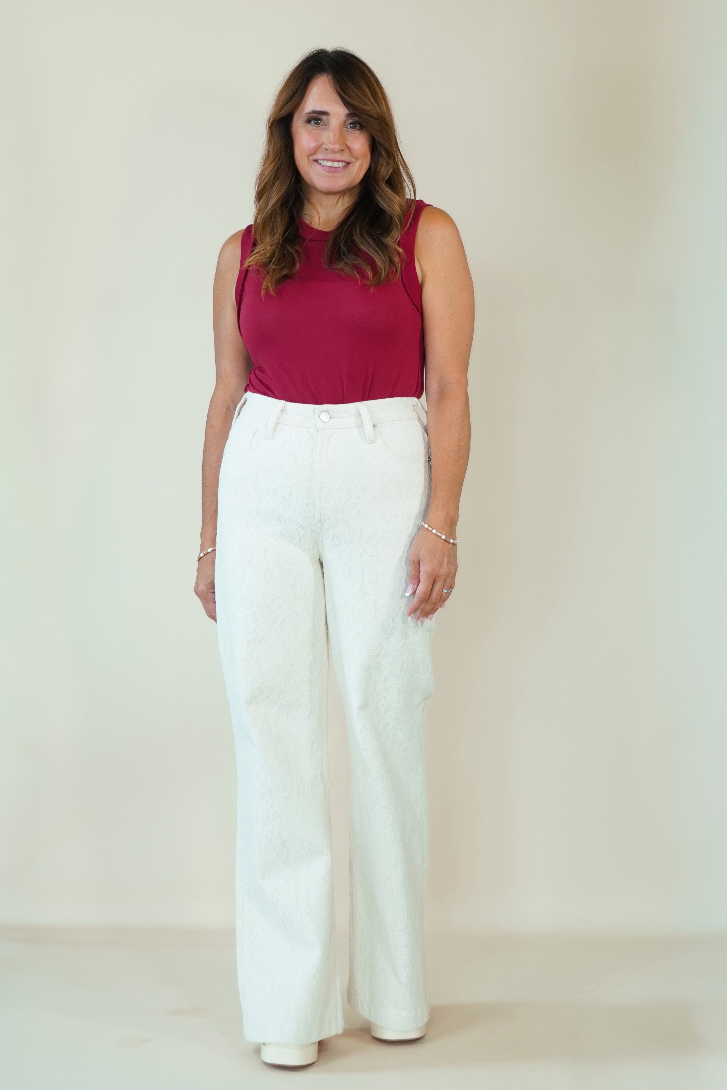 A woman modeling a sleeveless burgundy top and high-waisted white wide-leg pants against a plain beige studio background.