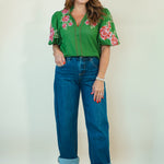 A woman modeling a green short-sleeve blouse with red and pink floral embroidery on the sleeves and collar, paired with blue jeans and beige heeled sandals, against a plain off-white studio background.