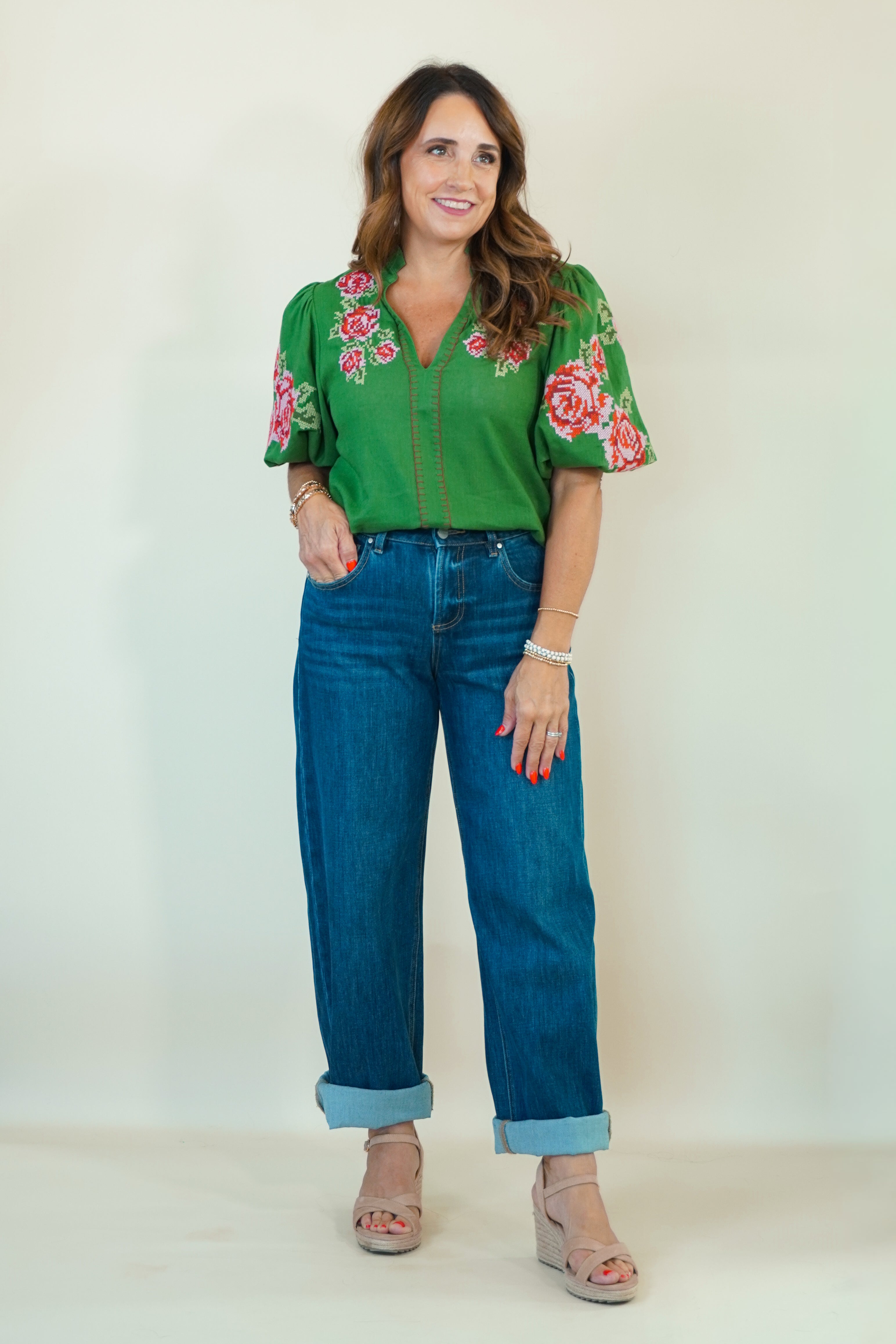 A woman modeling a green short-sleeve blouse with red and pink floral embroidery on the sleeves and collar, paired with blue jeans and beige heeled sandals, against a plain off-white studio background.