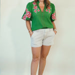 A woman modeling a green short-sleeve blouse with pink and red floral embroidery on the shoulders and sleeves, paired with white shorts and brown slide sandals, standing against a plain off-white background.